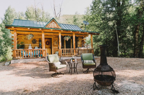 A cozy log cabin surrounded by trees, featuring a porch, outdoor seating, and a fire pit in front.